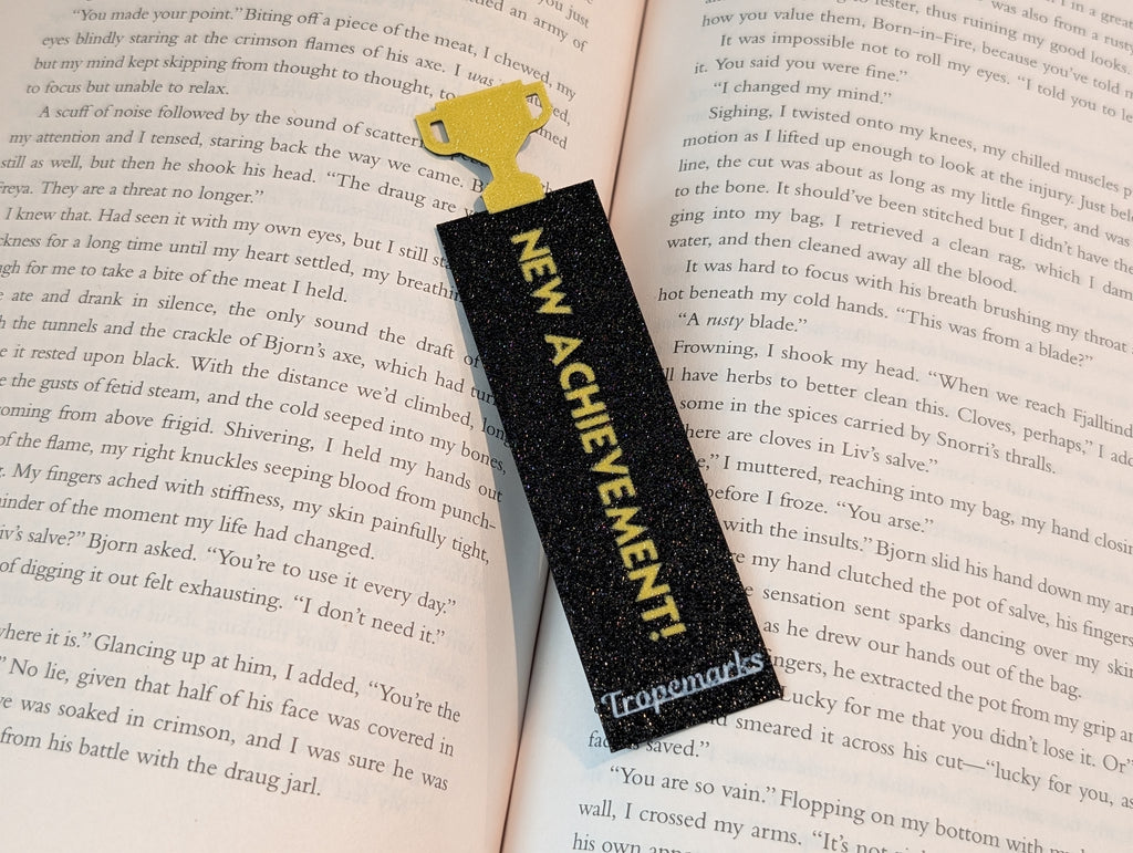 Black rectangle bookmark with yellow text reading "NEW ACHIEVEMENT!" with a yellow trophy icon on one end