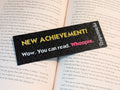 Black rectangle bookmark with yellow text reading "NEW ACHIEVEMENT!". Text below in white reading "Wow. You can read.". Magenta text reads "Whoopie."