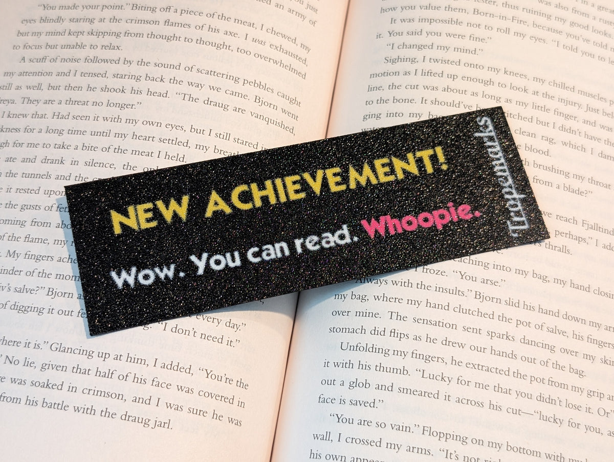 Black rectangle bookmark with yellow text reading "NEW ACHIEVEMENT!". Text below in white reading "Wow. You can read.". Magenta text reads "Whoopie."