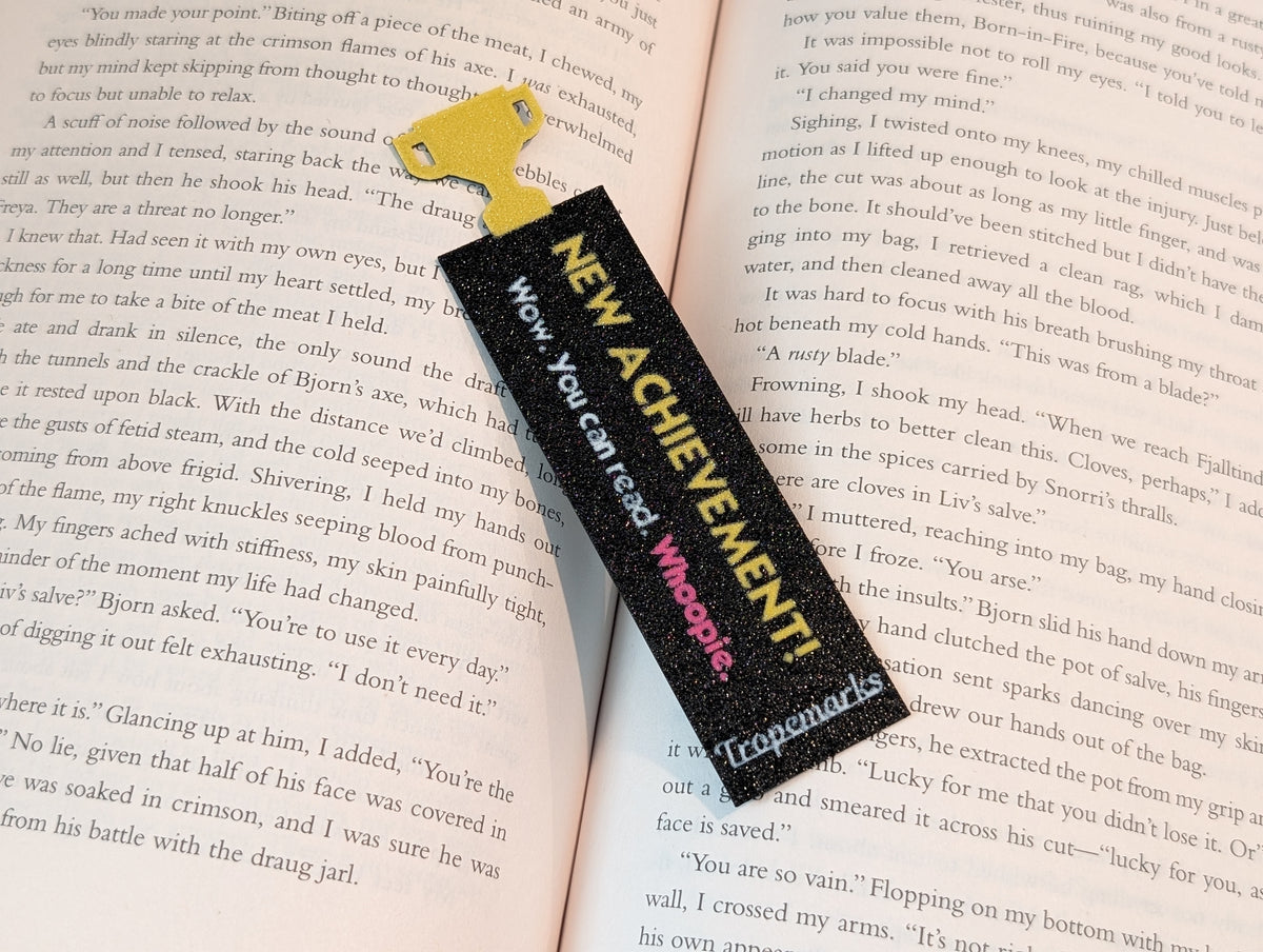 Black rectangle bookmark with a yellow trophy icon on one end. Yellow text on the bookmark reads "NEW ACHIEVEMENT!". Text below in white reading "Wow. You can read.". Magenta text reads "Whoopie."