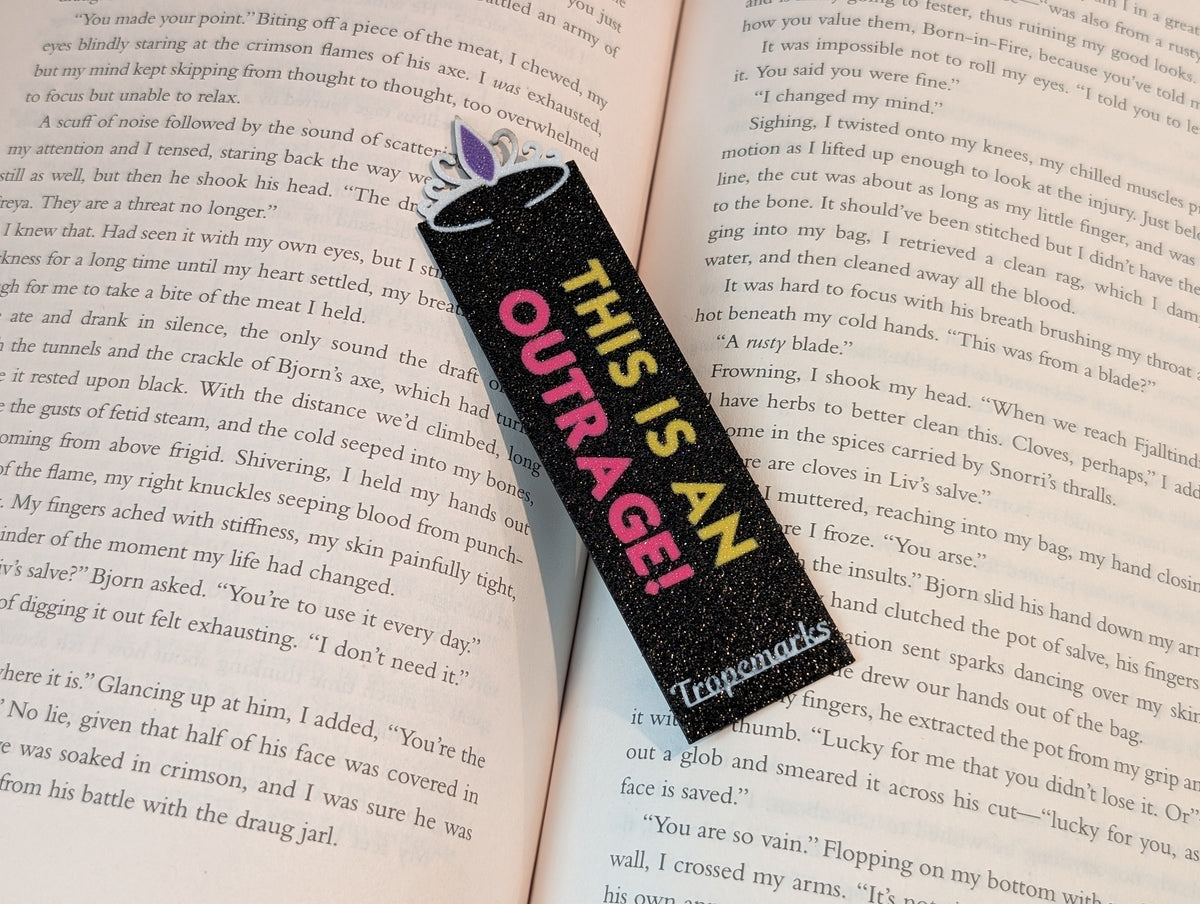 Black rectangle bookmark with a tiara icon on the end with a purple jewel in the middle. Text on the bookmark reads "THIS IS AN" in yellow and "OUTRAGE!" in magenta.